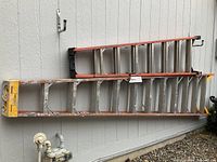 Two aluminum step ladders, 12 ft and 8 ft, laid horizontally against an exterior wall, orange rails with paint splatters and yellow ladder feet visible.