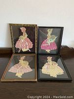 Four framed artworks showing Victorian women in full dress made from lace and ribbons, set on a cabinet.
