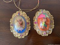 Front view of two vintage Liddle Kiddle Lucky Locket dolls Luana and Lola in ornate gold-tone lockets with pink and white decorative accents, showing clear plastic domes and dolls inside.