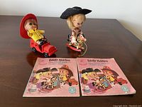 Overview and close-up of two Liddle Kiddles dolls Benson Burnie & Calamity Jiddle with their booklets and some accessories on a wooden table.