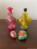 All four items displayed on a wooden surface showing the pink and yellow cologne bottles with ring and brooch in front.