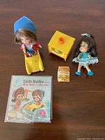 Two Liddle Kiddles dolls Millie Middle and Babe Biddle with yellow car, TV toy, miniature pretzels box and booklet displayed on table.