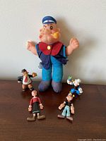 Five figures including a large fabric Popeye and four smaller plastic figurines representing Popeye characters, displayed on dark wood surface.