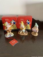 Three Royal Doulton Snow White and the Seven Dwarfs figurines with two original red boxes featuring Snow White illustration behind them on table surface.