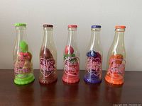 Five Soda Pop Kiddles dolls inside clear plastic soda bottle cases arranged side by side: Luscious Lime, Kleo Kola, Shirley Strawberry, Greta Grape, Olivia Orange, on wooden surface.