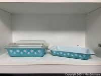 Two light blue Pyrex rectangular baking dishes with white snowflake patterns, one stacked with a matching clear glass lid on top, shown on a white shelf.