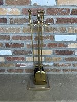 Full view of the vintage brass fireplace tool set including brush, shovel, poker, and stand in front of brick wall.