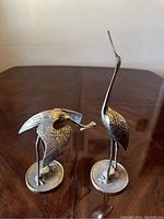 Pair of two brass heron sculptures on a wooden table, one standing upright and the other bending forward.
