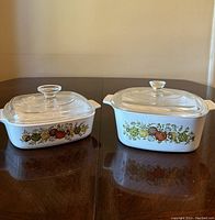 Two vintage Corningware rectangular baking dishes with vegetable pattern and clear glass lids on a wooden table.