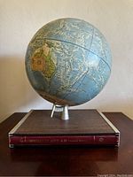 Photo showing the globe mounted on a small metal tripod and resting on a World Atlas book, displaying Pacific and surrounding areas.