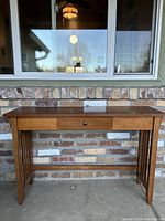 Front view of the vintage Mission style desk against a brick wall, showcasing the single center drawer with black knob and vertical slat sides.