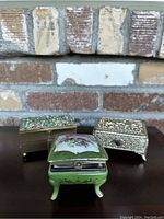 Green porcelain box with floral painting positioned in front, silver-tone ornate box, and brass-tone box with turquoise stones on a wooden surface against a brick background.