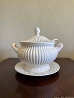 Front view of vintage white ceramic bowl with ribbed body, lid with knob, two handles, matching saucer, and spoon in lid slot.