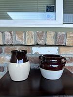 Two brown and white glazed stoneware pots against a brick background, one pitcher style and one two-handled pot.
