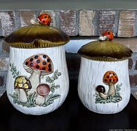 Full view of both ceramic mushroom canisters side-by-side showing colorful mushroom designs and ladybug handles on brown mushroom cap lids