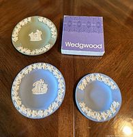 Three Wedgwood ceramic plates and the accompanying Wedgwood box arranged on a wood table.