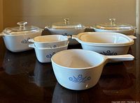 Full set of six vintage CorningWare baking dishes with Blue Cornflower pattern arranged on table. Three pieces have glass lids with glass knobs.