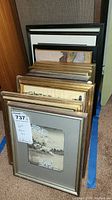 Stack of 15 framed artworks showing different frame finishes and thicknesses, predominantly wooden frames with gold and black colors.