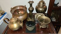 Overall view of brass items including tea pot, hanging cauldron, candlestick, bowls, vase, and others on table.