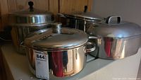 Four cookware pots including stock pots and roasting pan with lids on white surface against wooden background.