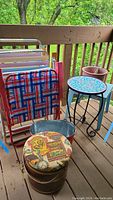 View of lot on wooden deck showing two metal folding chairs, metal Coca Cola tub, wooden bucket with upholstered lid, blue plastic table, black wrought metal mosaic top table, and planter