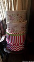 Photo showing four decorative round hat boxes stacked: two floral patterned with pink and cream flowers, one red and white vertical striped, and one red and white chevron patterned box.