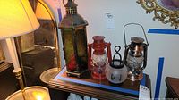 Wide shot of all four lanterns arranged on a wooden surface, showing the tall multicolored glass lantern, two smaller hurricane lanterns (red and silver), and the frosted glass candle holder with black deer motif.