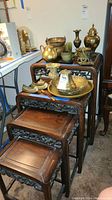 Set of four nested wooden tables with brass and ceramic decorative items on top, showing the tables' carved sides and rectangular tops.