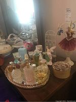 Perfume bottles arranged on a mirrored round tray with a pig-shaped item, white wire basket, fairy figurine, and decorative box on wooden surface.