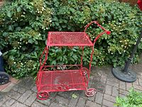 Red metal two-level bar cart shown outdoors on stone patio with green shrubbery background.
