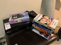 Sony AVCHD Blu-ray/DVD player, Casio TV-2000 LCD mini TV box, remote control, and stacked VHS/DVD tapes on black surface