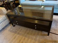 Wood coffee table with clear beveled glass top, four drawers with round metal pulls, dark wood finish, on carpeted floor between sofas.