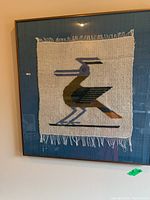 Framed woven textile wall art with bird motif displayed on wall.