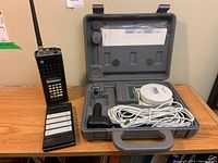 Wide view showing Sony GPS system in gray hard case with accessories and YAESU Triband handheld marine radio with keypad standing beside it on table.