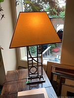 Lit table lamp showing metal geometric base and beige cloth shade near window.