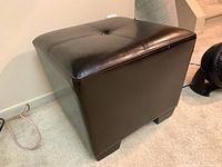 Angled side view of the dark brown faux leather ottoman showing the button tufted top and tapered sides.