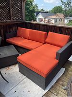 Two-piece woven patio couch set with bright orange cushions, situated on a wooden veranda with wooden railings.