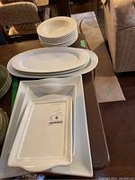 Stack of round white ceramic soup bowls with raised dot rim pattern on top of a collection of oval and rectangular white platters and plates.