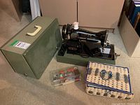 Full lot showing Singer sewing machine, green carrying case, plastic box with sewing supplies, and woven sewing box on carpet.