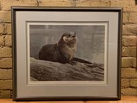 Full view of framed print showing otter on log with gray matte and wood frame against brick wall.