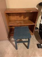 Wood shelving unit with two shelves and a blue painted wooden side table placed in front on carpeted floor.