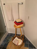 Photo showing yellow wooden stool with stacked assorted towels on top, white towels hanging on wall rack, wicker basket on floor with towels inside, and corner of tiled floor and wall.