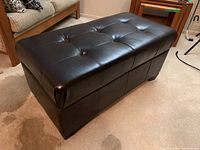 Angled front and side view of the black vinyl storage ottoman showing tufted cushioned top and block legs.