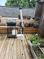 Full view of the Tasco telescope mounted on tripod outside on wooden deck with plants and barbecue in background.