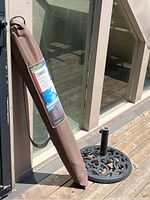 Patio umbrella in mocha brown case leaning against a structure next to a black metal umbrella stand with a decorative pattern.