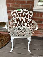 White cast aluminum patio chair with ornate curved back and floral cut-out seat, shown front view against brick wall background on a porch deck.