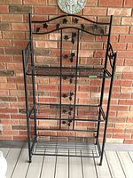Front view of black steel bakers rack against brick wall showing grapevine decorative elements and three collapsible shelves.