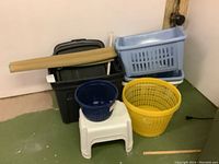 Photo showing all items: plastic step stool, assorted plastic laundry baskets in blue, yellow, and dark blue, stacked storage containers in black and white with lids, toilet plunger with white handle, and rolled bamboo style window shade on top.