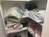 View of two shelves with various folded blankets, comforters, and bedspreads including beige comforter in packaging, various folded linens, and white comforter with maroon trim.