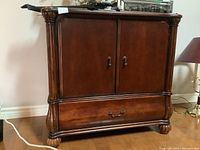Front view of wooden cabinet showing two doors and single drawer with brass handles, dark wood finish, and carved details on legs.